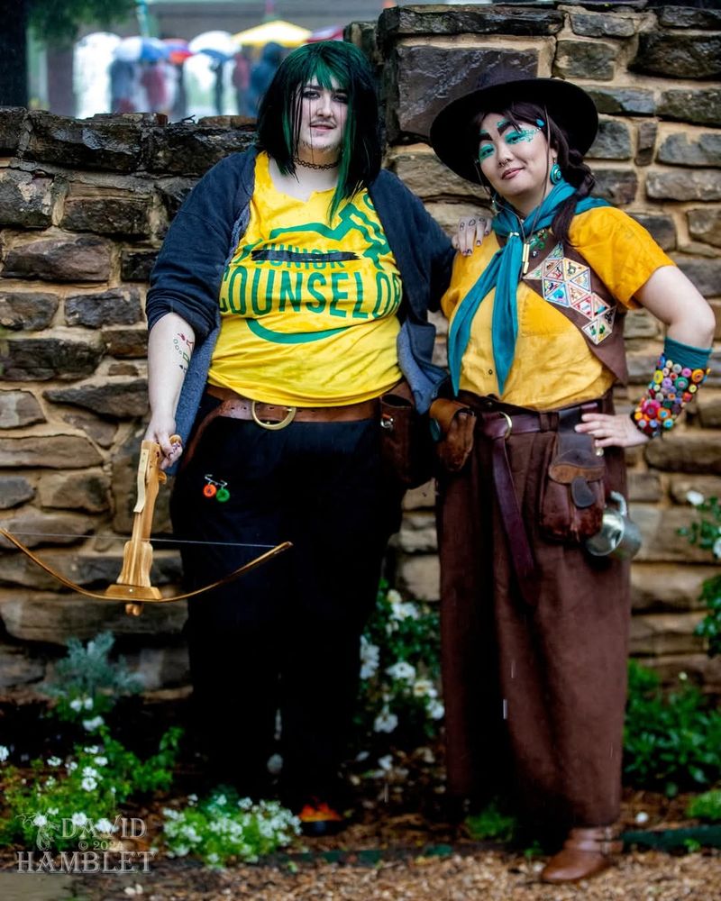 Photo taken by David Hamblet

Purpledragon is on the left cosplaying Luc Brenatto & JWo on the right cosplaying Veth Brenatto. Luc Brenatto is wearing a yellow t-shirt with the words "Junior Counselor" but the word "Junior" is marked through in black. They are also holding a wooden crossbow in their right hand. Veth Brenatto is wearing a mustard yellow blouse with a scout badge sash, brown hat, teal neckerchief, & button adorned arm guards.