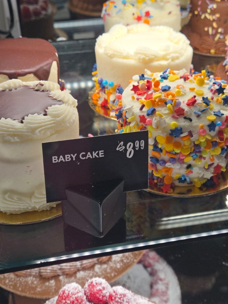 Photo of a bakery display with mini cakes and a black standee sign in front of the cakes reading "Baby Cake $8.99"