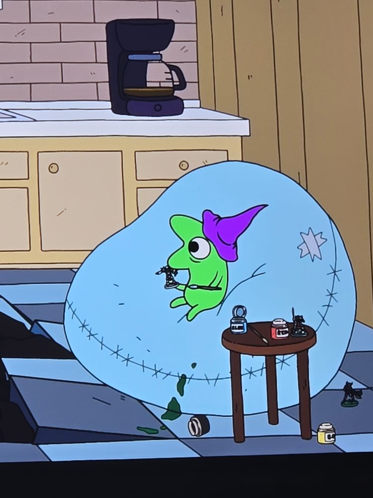 Glep, a small lime green cartoon character from animated series Smiling Friends, sits on a pastel blue bean bag holding a Warhammer 40k miniature figure in one hand and a paintbrush in their other hand with a shocked expression on their face. There is a a brown, presumably wood, round side table nearby holding 2 paint cans colored blue and red, a paintbrush, and Warhammer 40k miniatures. There is a side turned green pain container with spilled green paint on the bean bag. A yellow container of paint sits by the back leg of the side table and a small unpainted figurine on the blue on blue toned large checkered flooring. In the background are light tan cabinets with a red brick backsplash and a black coffee maker with a mostly empty coffee carafe. 