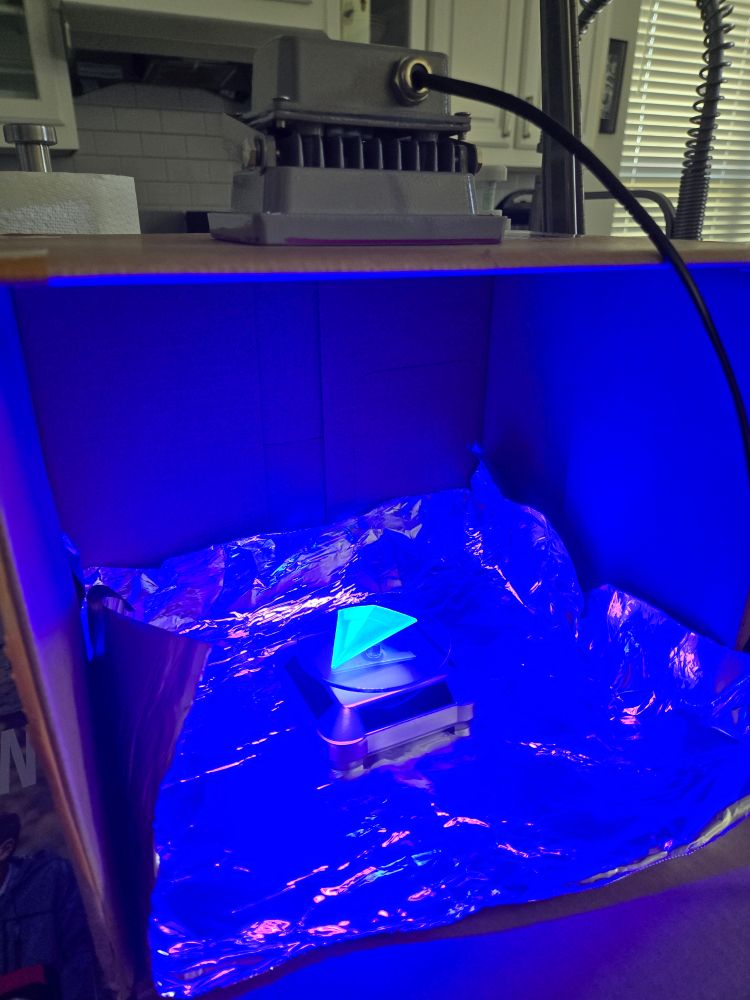 Triangular resin printed piece glows bright neon green under a UV light in a box surrounded in silver foil and sitting on a solar rotating plate.