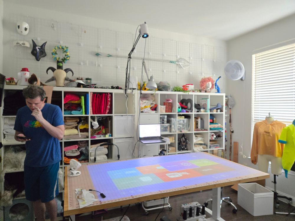 Photo of JWo's craft room. In the middle is a large cutting table with a gridded cutting mat on top with a operating menu projected onto the table from above from a white, small projector mounted on an adjustable mic arm. To the left of the photo is Drift0r, wearing a blue gay frogs shirt with darker blue shorts, and seemingly picking his nose & looking down at his phone in his hand.
