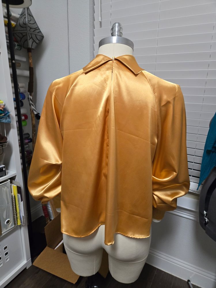 The back view of a golden satin blouse with billowing high cuffed long bell sleeves and foldover collar neckline. It is worn on a ivory canvas sewing mannequin.