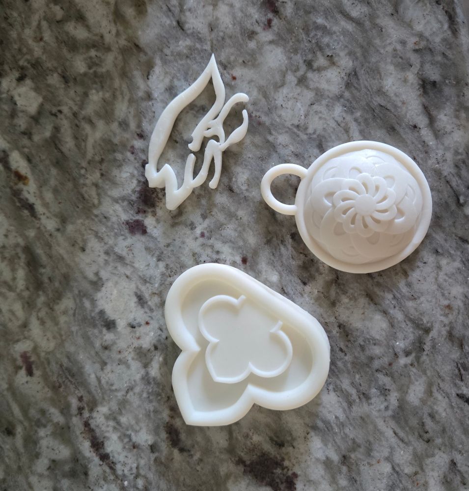 3 white resin printed small pieces. Top piece is an abstract shape, middle piece is a circular pendant with a floral motif, and bottom piece is a clover shaped piece.