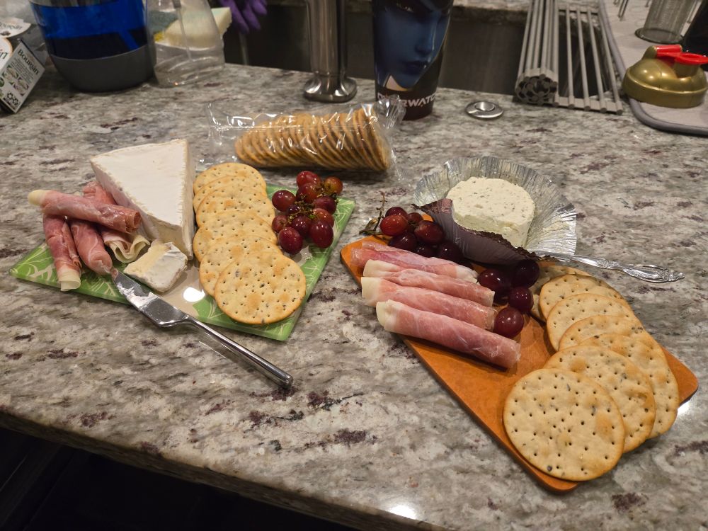 Two charcuterie boards side by side on a grey speckled granite countertop. The left board has brie, prosciutto & mozzarella rolls with red grapes and olive oil and rosemary crackers. The right board has all the same sides, except the brie has been replaced with a Boursin brand black truffle & sea salt soft, spreadable cheese
