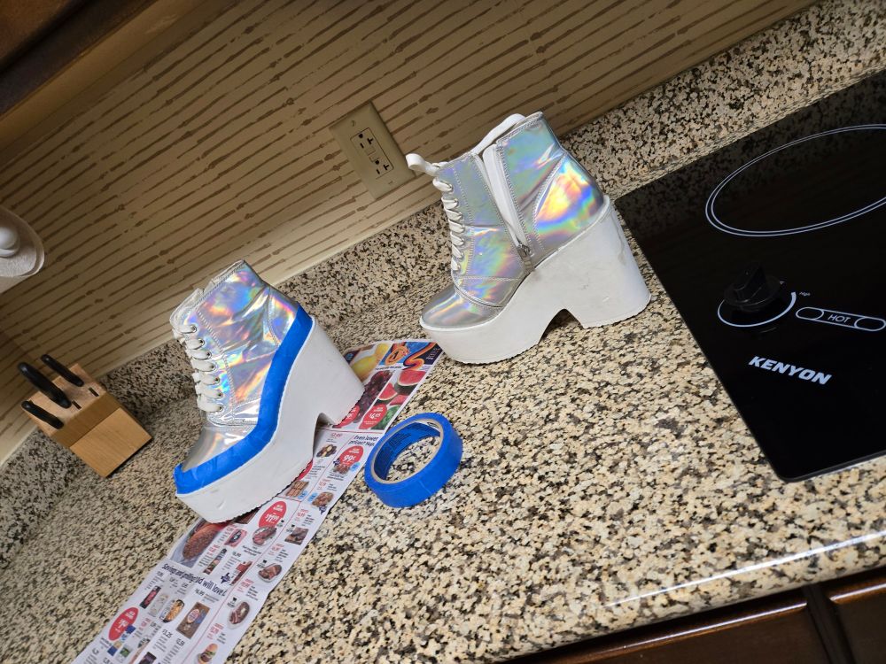 A pair of iridescent silver platform ankle boots with white platform bases sit on a speckled tan granite kitchen counter top. The boot on the left has blue painter's tape wrap around the base and sits on top of a newspaper grocery ad. A roll of blue painter's tape sits between the boots. 