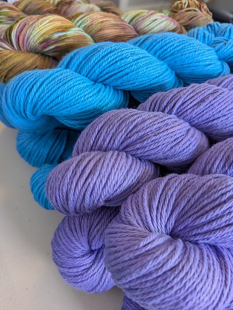 Purple Wisteria yarn and bright blue Blue Poppy yarn, with Cafe Deluxe DK in the background - a variegated color way with tan, blue, yellow, and mauve.