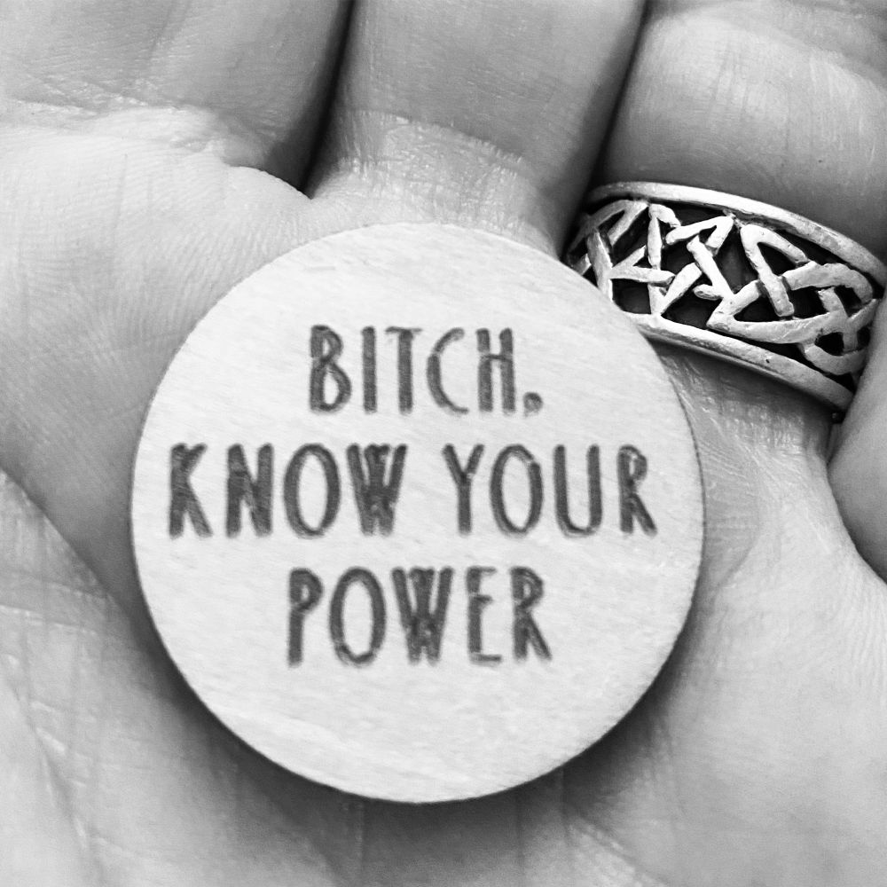 Black and white photo of a hand holding a round disk with the words Bitch Know Your Power on it. Celtic knot ring on a finger in the background. 