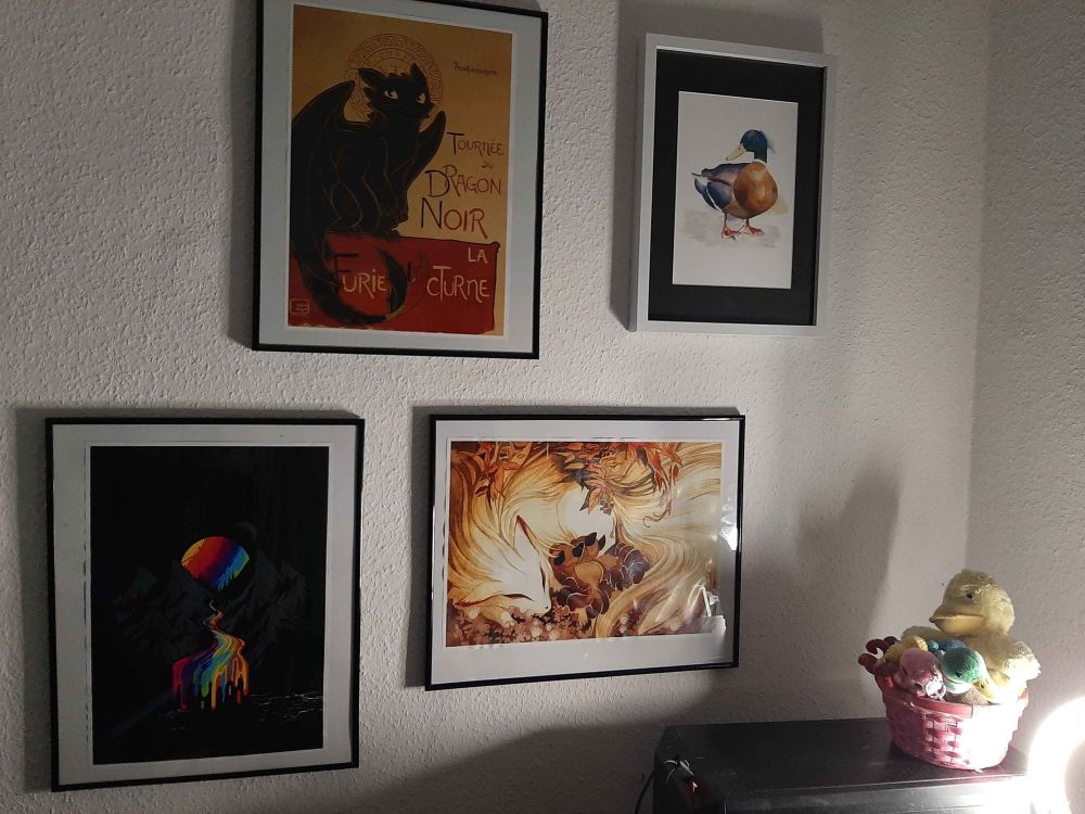 A photo of a few paintings hung on a wall next to a black PC tower. the two top paintings show toothless, one of the main dragons from "How to train a dragon", depicted like the famous poster "Tournee du chat noir" and a painting of a mallard. The two lower paintings are a mountain range with a rainbow colored sun behind them that seems to melt into a rainbow colored river flowing to the front of the painting and a mystical fox demon feeding her pups that are snuggled up to her.

On the PC is a pink basked filled with plushies, which are all ducks and octopusses
