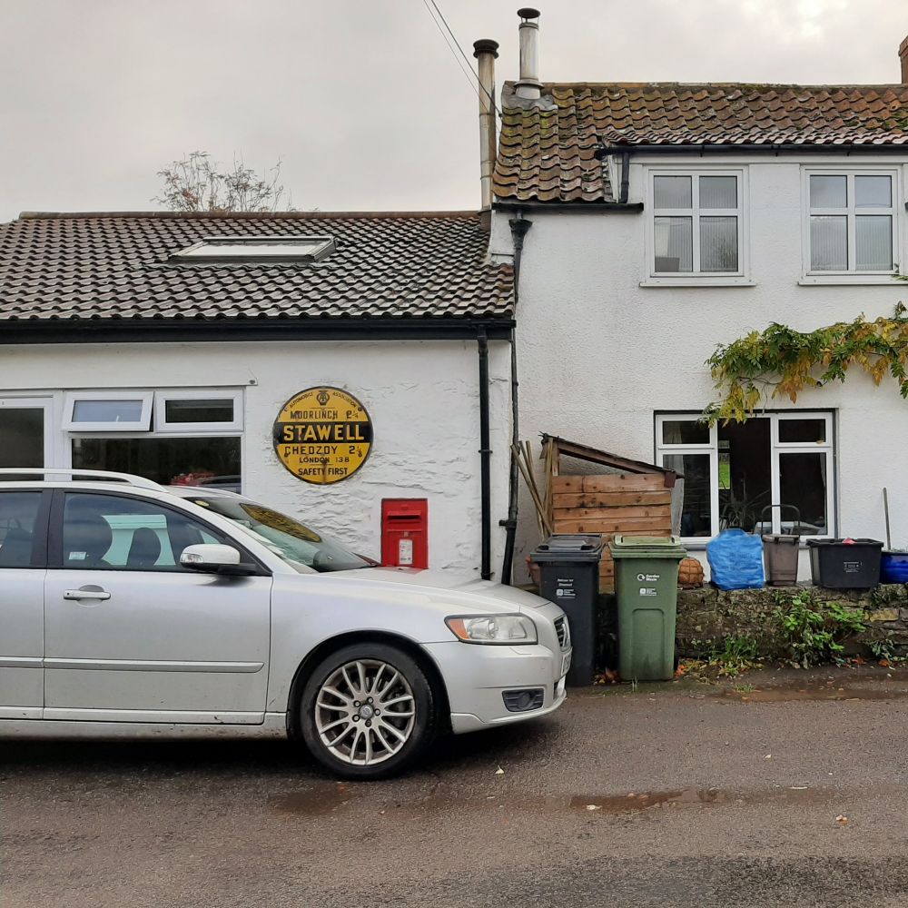 Old AA roadsign in the village of Stawell. It's a yellow metal plaque on the wall with black writing. There's a car in front of the wall (naturally) and a red wall postbox.