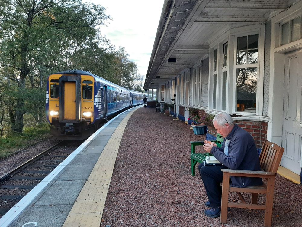 View along the platform with part of the station hostel included. A train is approaching, and a man sits on a bench checking his phone.
