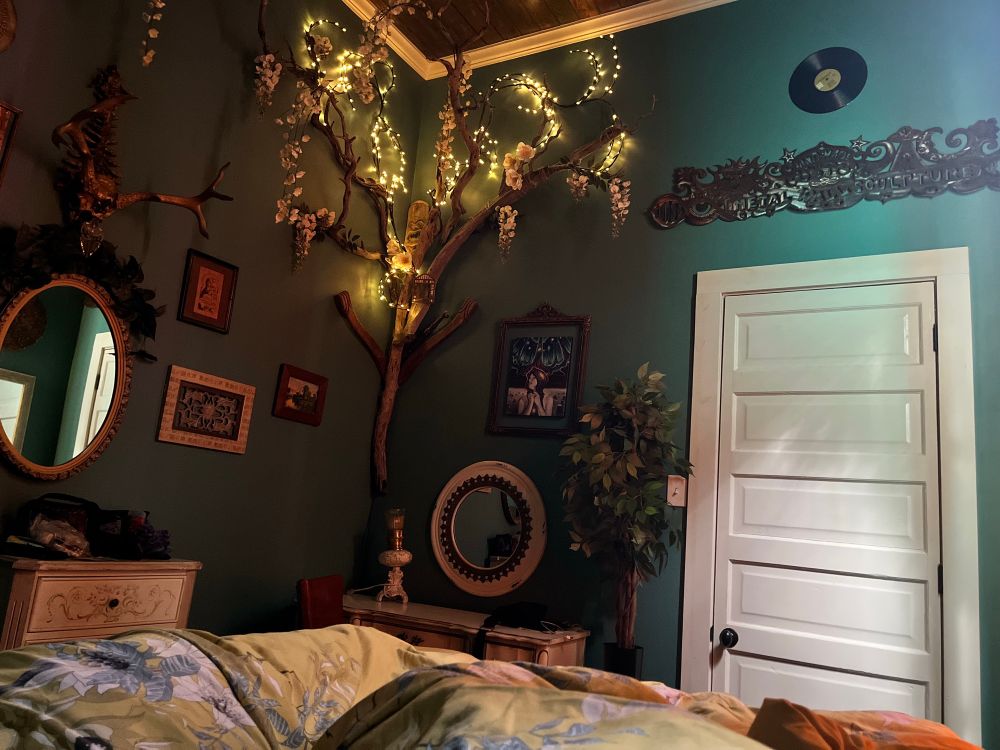 View of a small bedroom from the bed. It has high ceilings and teal walls with driftwood illuminated by fairy lights hanging in one corner. A tiny desk sits underneath. The comforter is large and soft and has flowers and birds printed on it.