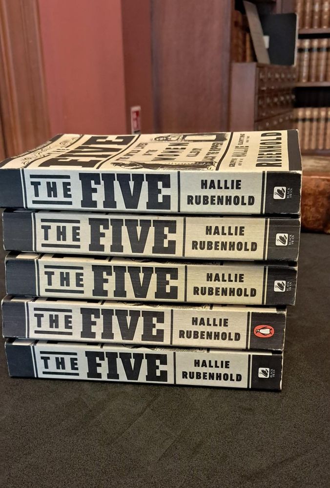 Five copies of 'The Five' by Hallie Rubenhold sitting on a table with library shelves in the background. 