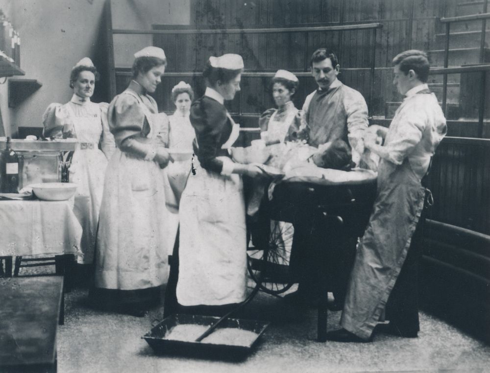 Black and white photograph of an operating theatre with a surgeon, assistant and team of nurses looking over a patient on the table. The men are wearing aprons and there is an empty lecture theatre behind them.