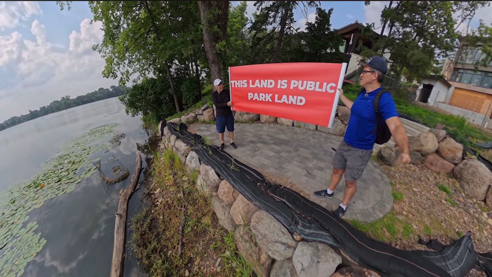 Larry Werner & Wedge Live’s John Edwards unfurling a “This Land is Park Land” banner on a garish stone patio encroaching on Minneapolis public land around Cedar Lake. Yes, the home in the background is a tear down. 