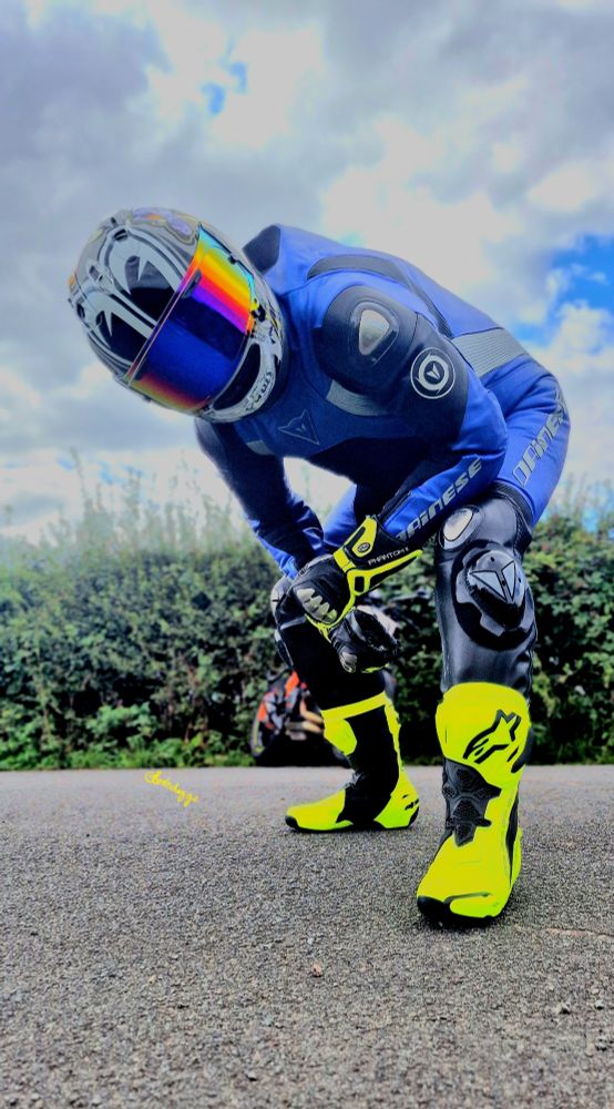 Man wearing a blue sports bike leather suit, yellow boots, yellow gloves and a multicoloured helmet and rainbow visor, bending over looking down at you