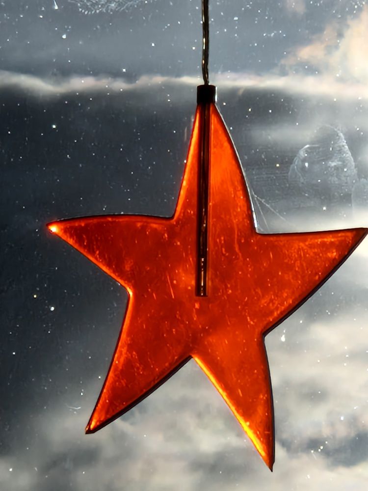 Ein roter Stern aus plexiglas vor Fenster mit winterlichen Wolken im Hintergrund