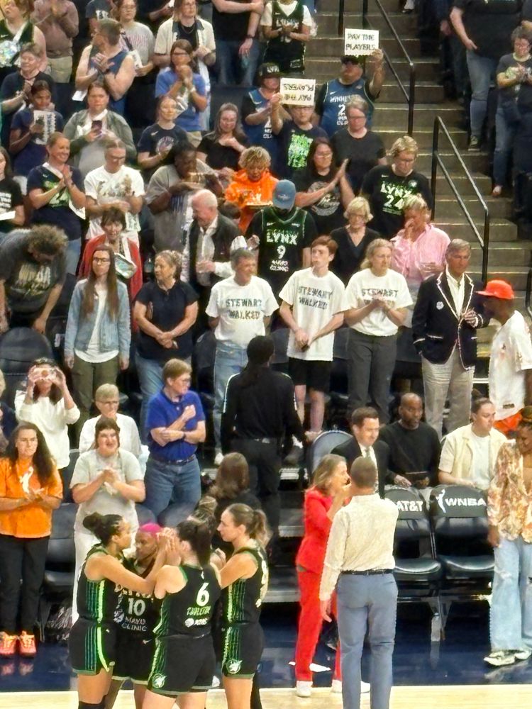 3 people in white t shirts in the stands of Target center that say “STEWART WALKED”