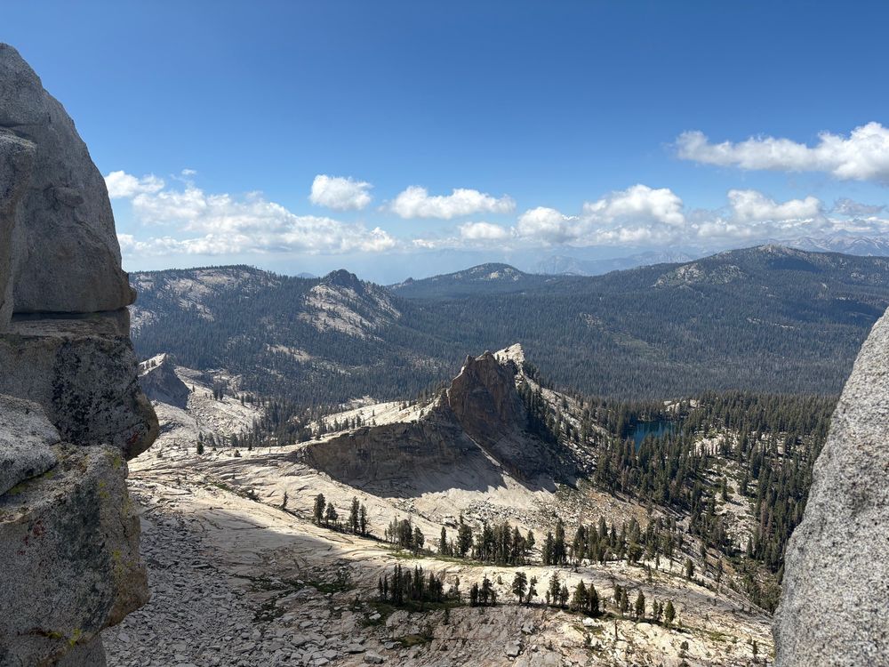 Photo of a very small alpine lake in the high sierras. There are many pine trees in the background and lots of granite in the foreground. The sky is a clear blue with some nice white poofy clouds 