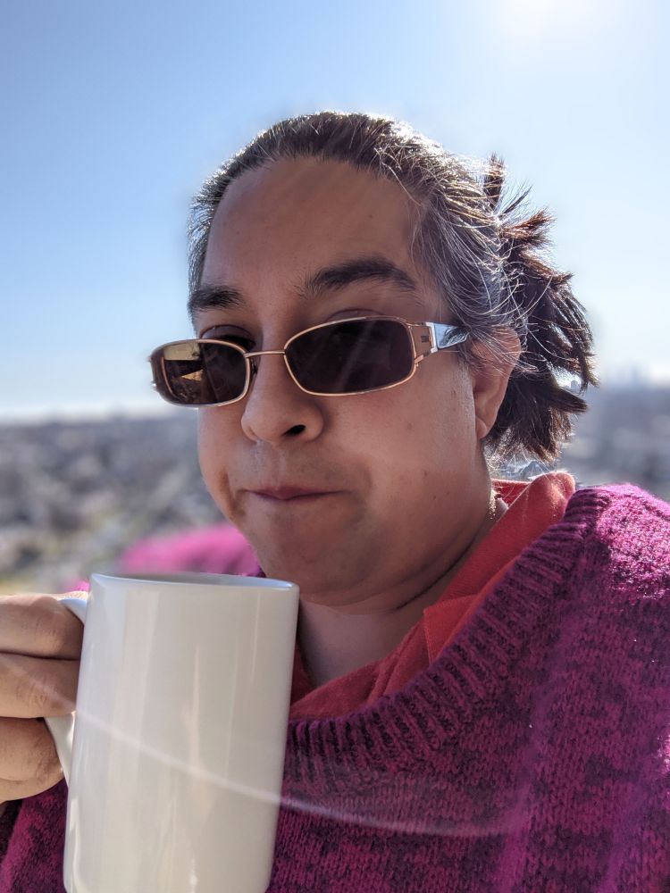 A brown-skinned woman with graying dark brown hair in a spiky bun, wearing a magenta cardigan and dark sunglasses enjoying a cup of tea in a white mug outside in the bright morning sun on a brisk winter day. 