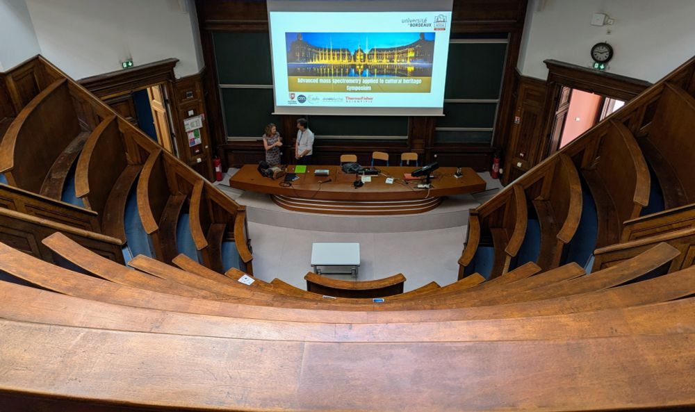 Bird's eye view of a circular lecture theatre with curved wooden tiered seating arranged in concentric arcs. At the bottom, a projection screen displays "Advanced mass spectrometry applied to cultural heritage Symposium" with University of Bordeaux branding. Two people stand near the presentation area at the front, with wooden chairs and desks arranged in the curved amphitheatre design typical of academic venues.