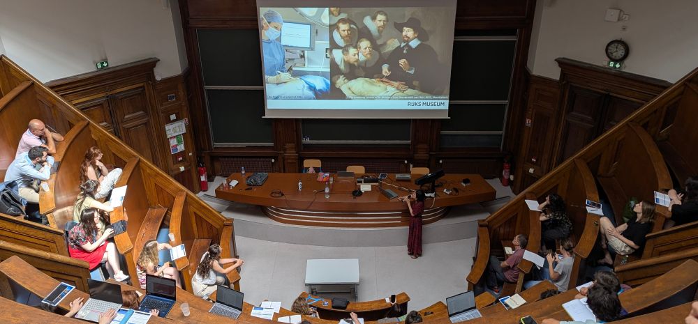 
A packed amphitheatre lecture hall with curved wooden tiered seating filled with attendees listening to a presentation. The projection screen displays a split image comparing modern mass spectrometry equipment on the left with Rembrandt's famous 1632 painting "The Anatomy Lesson of Dr. Nicolaes Tulp" on the right, branded with "RIJKS MUSEUM". The historic venue features elegant wooden architecture with multiple levels of curved benches rising steeply upward.