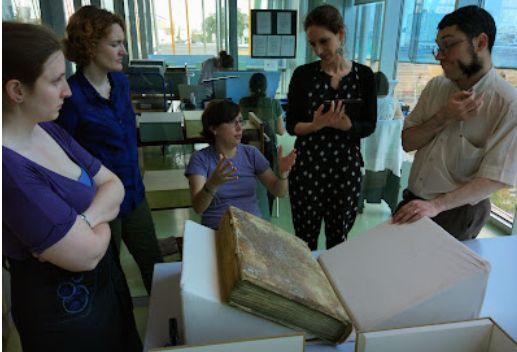 In 2016, the team in 2016 first confronted the collection at Troyes  - but what are the bindings made of?  Image of five people, one sitting, discussing a large bound medieval text held by supports in a modern library. 