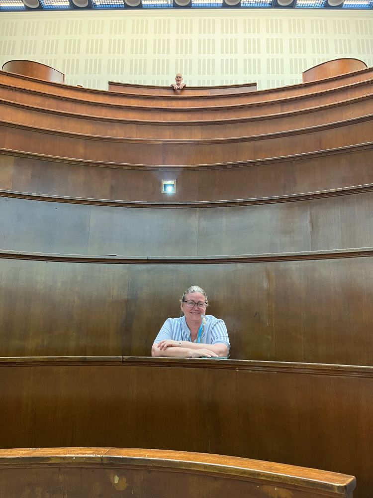 A woman with glasses and light blue striped shirt sits at a curved wooden desk in a tiered lecture theatre. The amphitheatre features multiple levels of curved wooden benches rising upward, with another person visible in the distant upper seating. The architectural design shows elegant curved lines and wood paneling throughout the academic venue.