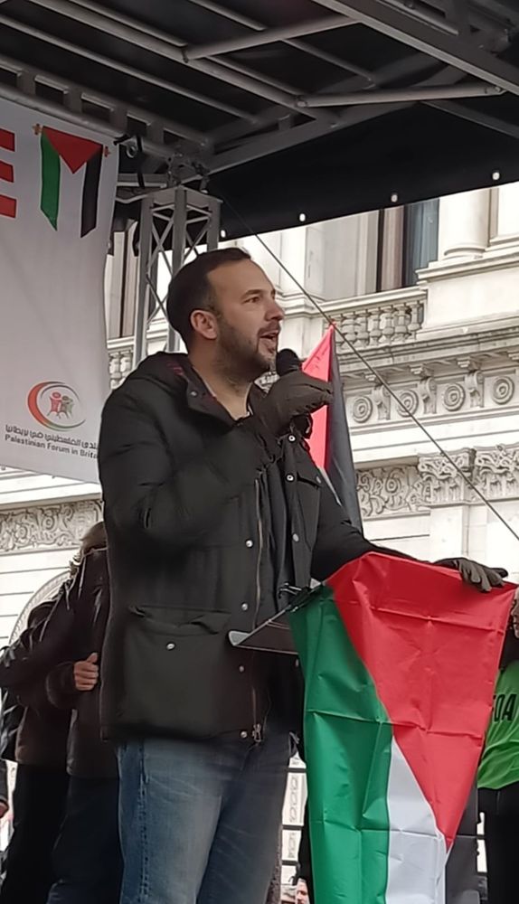 Zack at a podium behind a Palestinian flag.