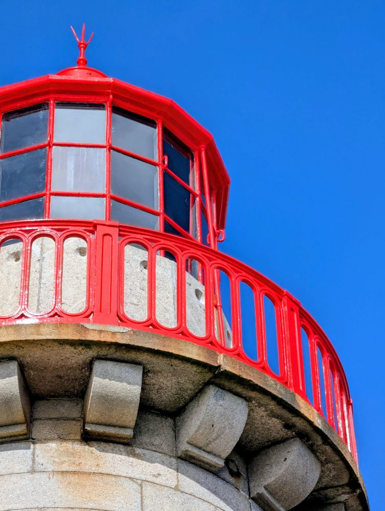 A close-up, low-angle shot of the top of a red and grey lighthouse against a vibrant, cloudless blue sky. The red metal railing and the glass-paned lantern room are prominent against the stone of the lighthouse tower.