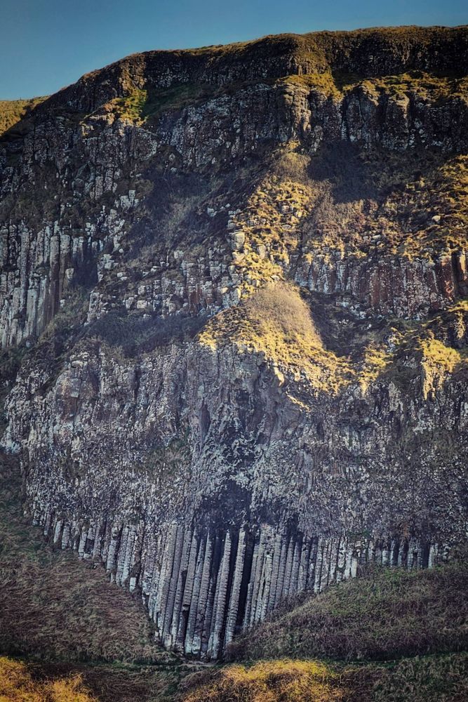 A close-up, sunlit shot of the rugged cliffs and striking basalt columns of the Giant's Causeway in Northern Ireland. The distinctive hexagonal columns rise from the base of the hill, their geometric shapes contrasting with the rough, mossy rock face above. The sunlight casts dramatic shadows, highlighting the texture and detail of the geological formation.