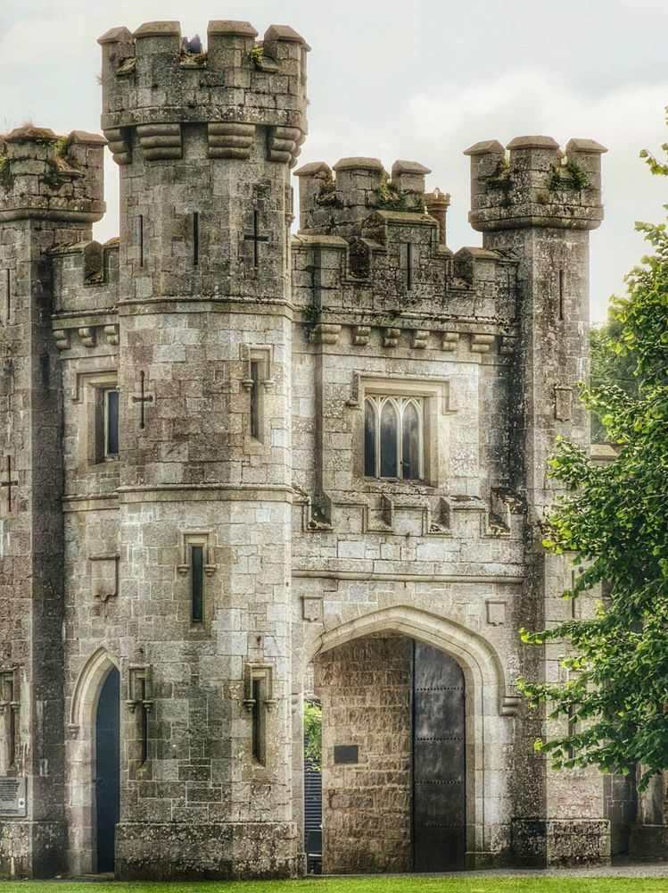 A close-up, eye-level shot of the grey stone entrance tower of a large, historic castle. The tower features several crenellated turrets, arched doorways, and rectangular windows with stone mullions. The stone blocks show signs of age, with some moss and discoloration. To the right, the vibrant green leaves of a tree are visible, adding a touch of nature to the ancient structure. The sky above is bright and slightly overcast.