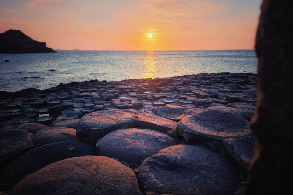 A wide shot of the Giant's Causeway in Northern Ireland at sunset. The sun, a bright orange orb, is low on the horizon, casting a golden path across the calm sea. The distinctive hexagonal basalt columns of the causeway fill the foreground and extend towards the water, bathed in the warm light of the setting sun. A dark silhouette of a cliff or headland is visible on the left side of the frame, and a partial, dark vertical element from the extreme right edge of the photo frames the scene. The sky transitions from a vibrant orange and pink near the sun to a softer blue higher up.