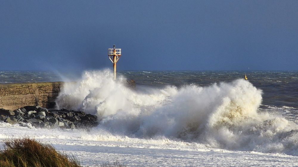 A dramatic photo of large white waves crashing against a stone sea wall on a stormy day. The sky is dark blue and grey, and the sea is turbulent. A red and white post with a light is visible on the sea wall and a yellow buoy is further out to sea. The foreground is a sandy beach with some dry grass and foliage.