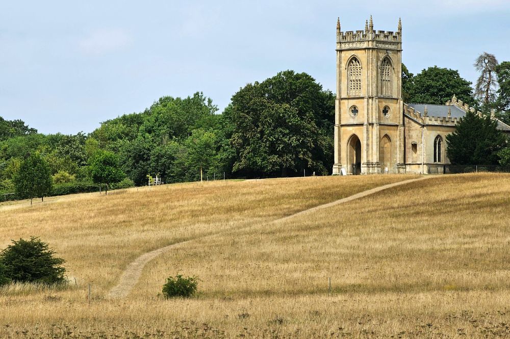 A lone, historic stone chapel with a tall steeple stands on a grassy hill, surrounded by lush green trees under a bright sky. A well-worn path snakes up the dry, golden-brown grass of the hill towards the chapel's arched entrance.