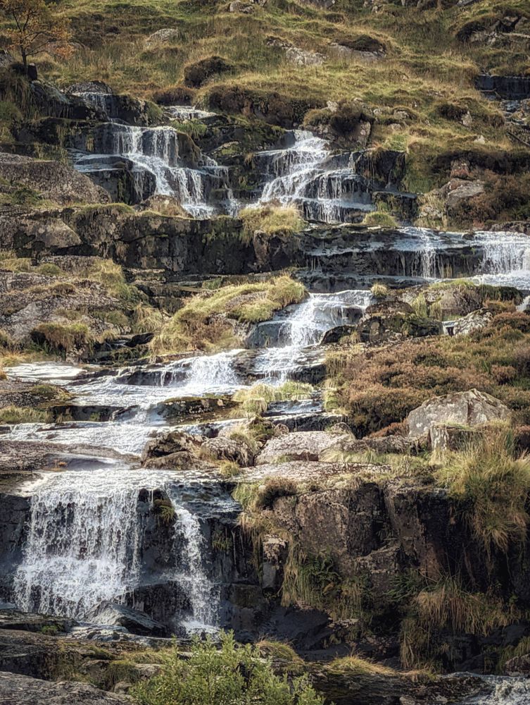 A vertical photograph of a multi-tiered, cascading waterfall flowing down a rocky, grassy hillside. The water rushes over numerous ledges, creating white foam and spray. The surrounding terrain is covered in rugged brown rocks and patches of dry, golden-brown moorland grasses and heather, suggesting a rural or mountainous area in autumn. The light highlights the flowing water against the darker, textured rock and vegetation.