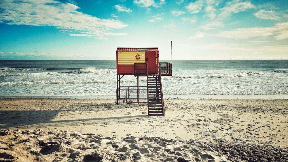 A slightly desaturated, wide-angle photo of a yellow and red lifeguard hut on stilts on a wide, sandy beach. A set of metal stairs leads up to the hut's door. The ocean is in the background with white waves rolling onto the shore under a blue sky with scattered white clouds. The overall tone is slightly retro and calm.