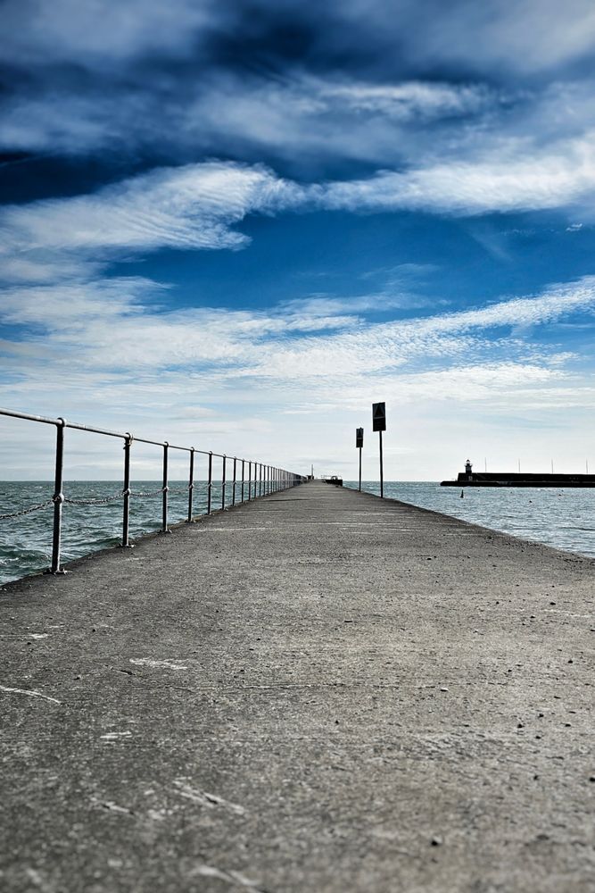 A long, concrete pier stretches into the distance under a dramatic blue sky with wispy white clouds. A metal railing lines the left side of the pier, separating it from the dark, choppy water. In the far distance on the right, a lighthouse is visible. The perspective is low, emphasising the length of the pier and the vastness of the sky and sea.