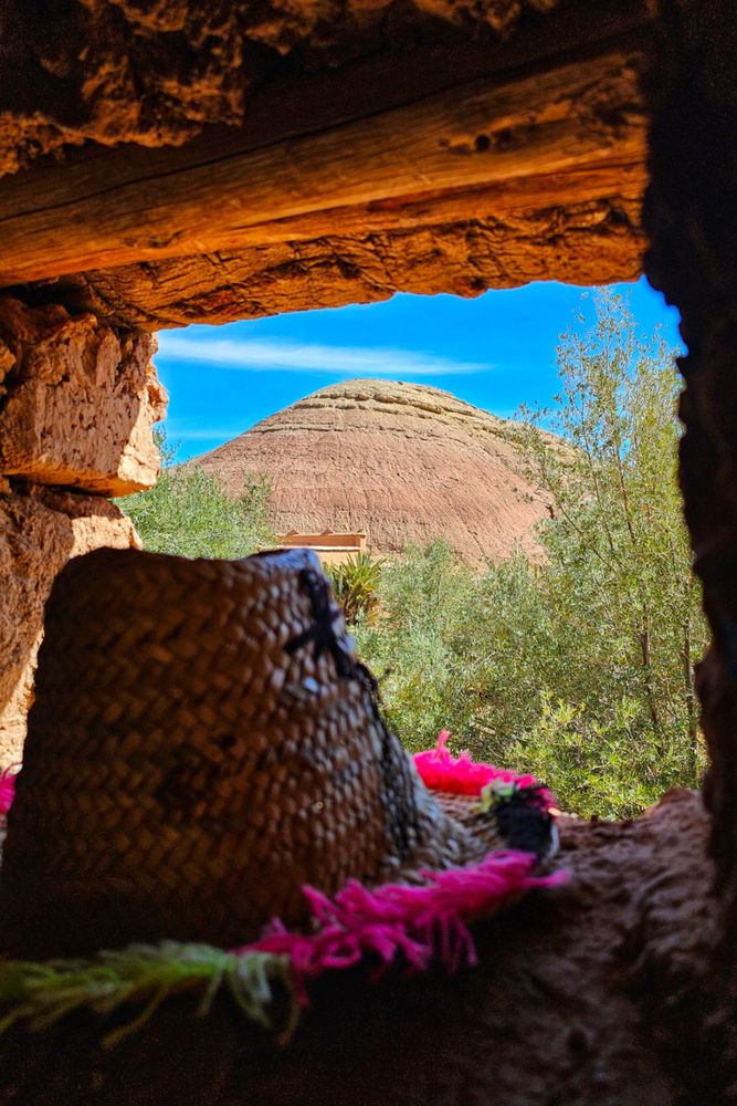 A close-up view through a rough wooden and stone window reveals a vast, reddish-brown, cone-shaped hill, part of the ancient Ksar of Ait Benhaddou, against a bright blue sky with a thin layer of clouds. In the foreground, out of focus, sits a woven straw hat with a colourful pink tassel, adding a touch of vibrant detail to the scene. Green shrubbery and trees fill the mid-ground between the window frame and the hill.