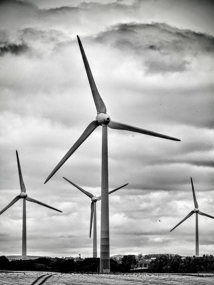 A high-contrast black and white photo of a field with four wind turbines silhouetted against a dramatic, cloudy sky. The turbines are in various sizes and distances, creating a sense of depth. In the foreground, the field is visible, and in the distance, a small building and some trees can be seen. The overall mood is dramatic and powerful.