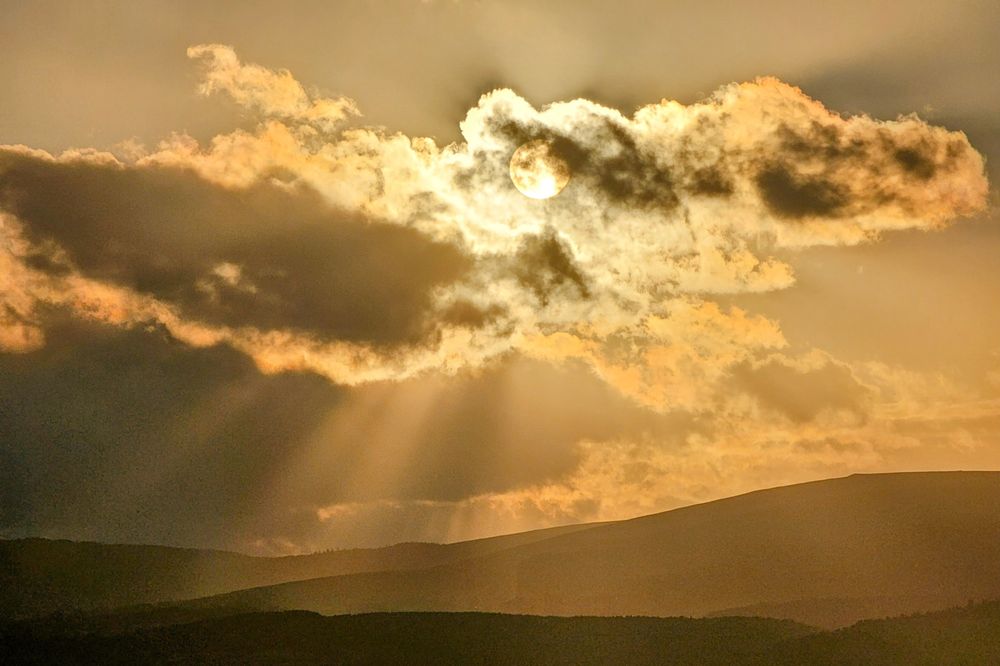 The sun, a bright circle, is partially obscured by a dramatic, textured cloud at the top of the frame. Golden sunbeams burst from behind the cloud, casting radiant light downwards onto a series of silhouetted rolling hills that stretch across the bottom of the image. The overall mood is warm, peaceful and a little hazy.