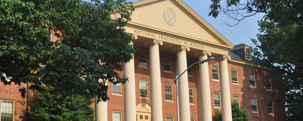 A red-brick building with tall white columns and a triangular pediment reading "National Institutes of Health," partially obscured by leafy trees.