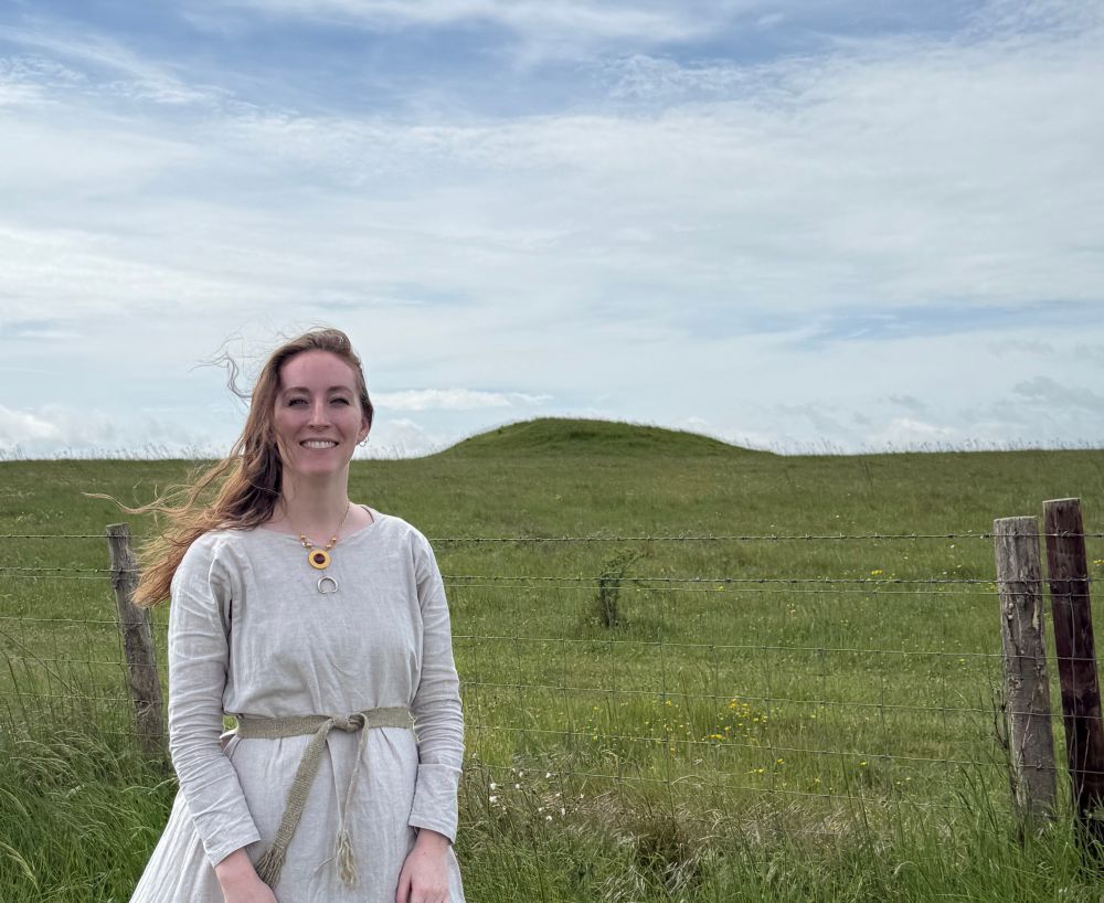 Woman standing outside the Bronze Age burial mound where the jewelry she is wearing replicas of was found. The jewelry is a pair of silver cow horns and a gold circular pendant with amber in the center. The woman is very happy and wearing an undyed linen dress.