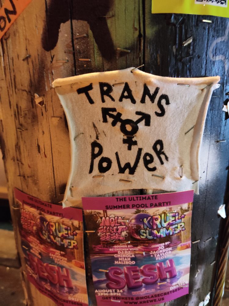 A piece of fabric stapled to a light post that says trans power