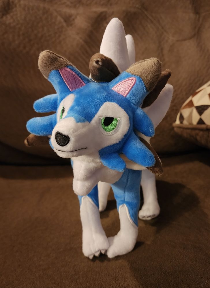 Photo of a poorly constructed bootleg shiny lycanroc plush with a silly expression and paws pointed together shyly