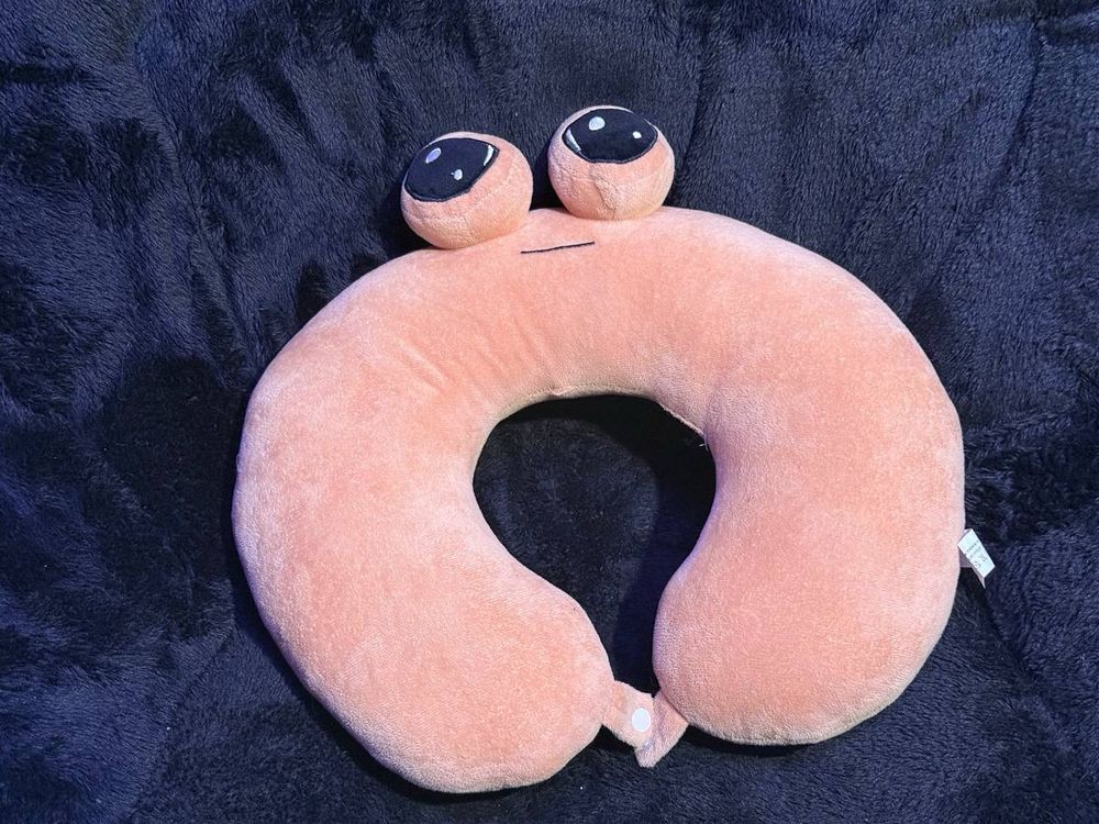Front view of a plush neck pillow with the characteristic pou expression and eyes.