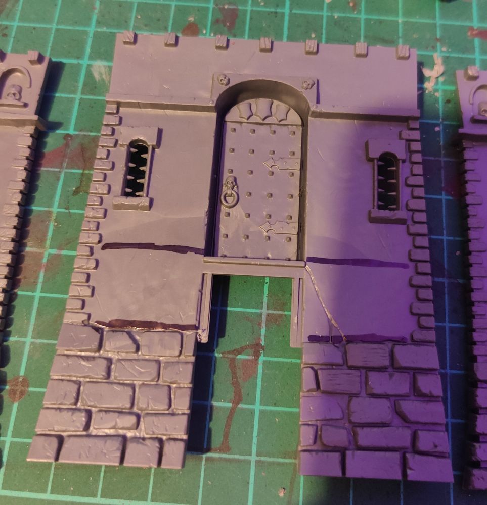 A single piece of the base of the tower. It is on a paint-stained green cutting mat. All pieces of the tower are in plain grey plastic. 
It has a black line drawn on it just above the bottom brick line and another just above the door stop.