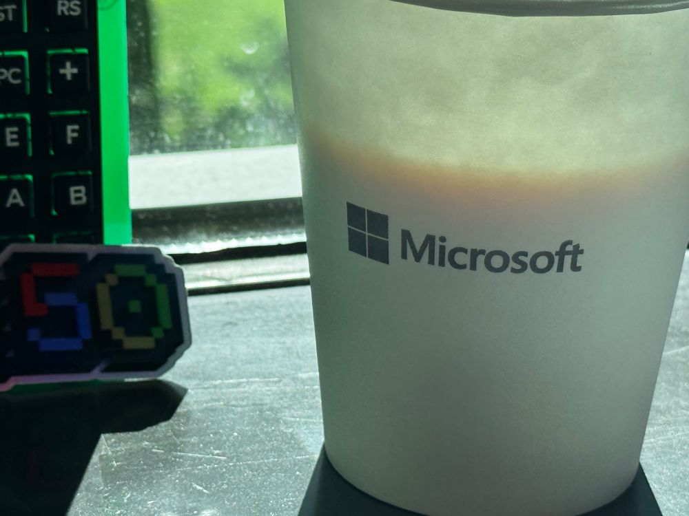 A Microsoft branded paper cup. Nerdy things available to the side: a 50th anniversary sticker and a circuit board and keypad. 