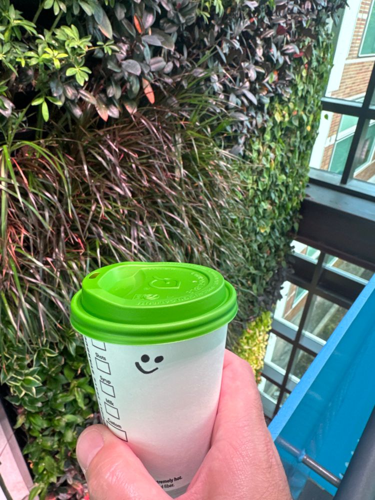 A latte  cup with a smile in front of a cool wall of plants. 