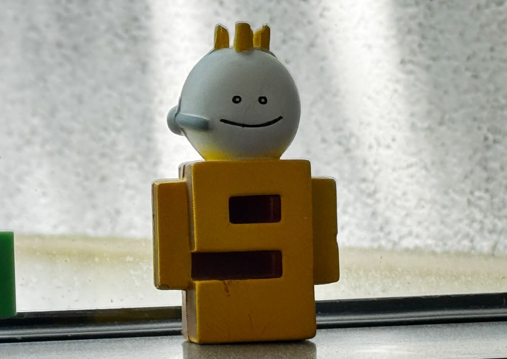 Small toy figure, a yellow stylized "9" with a spherical white ping-pong ball head wearing a headset.