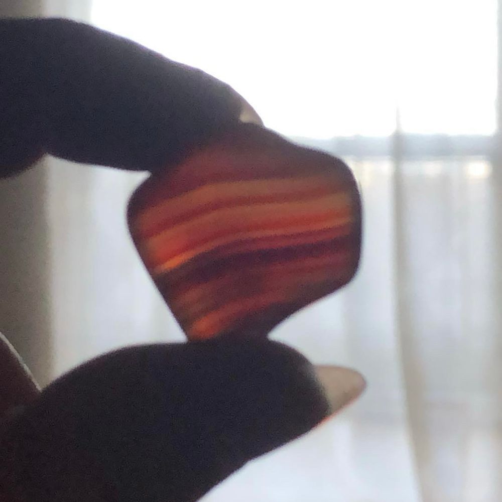 Closeup of someone holding a piece of carnelian between their finger and thumb. The crystal is backlit and orange and red stripes are clearly visible